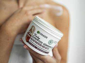 Tube der Creme von Garnier Body Superfood im Vordergrund, unscharf im Hintergrund eine Person, die mit der rechten Hand Creme auf Schulter oder Körper aufträgt.